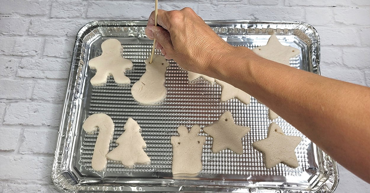 salt dough ornaments on a cookie sheet and a hand with a skewer