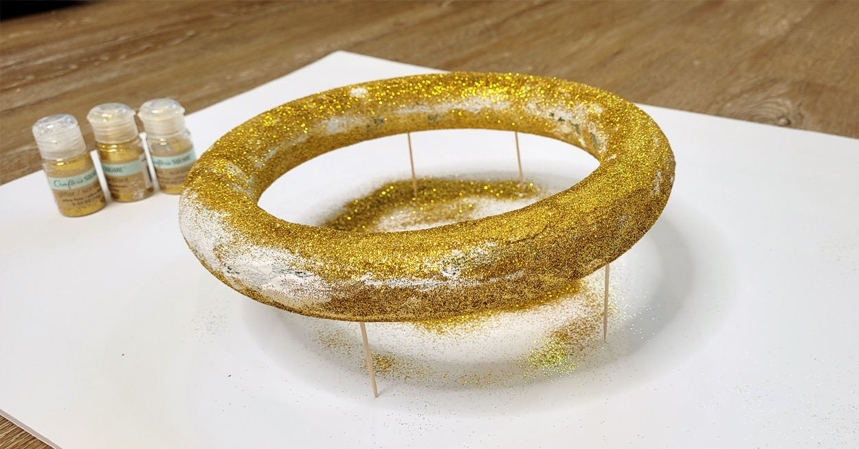 A foam wreath with toothpicks propping it up, that has gold glitter on it.