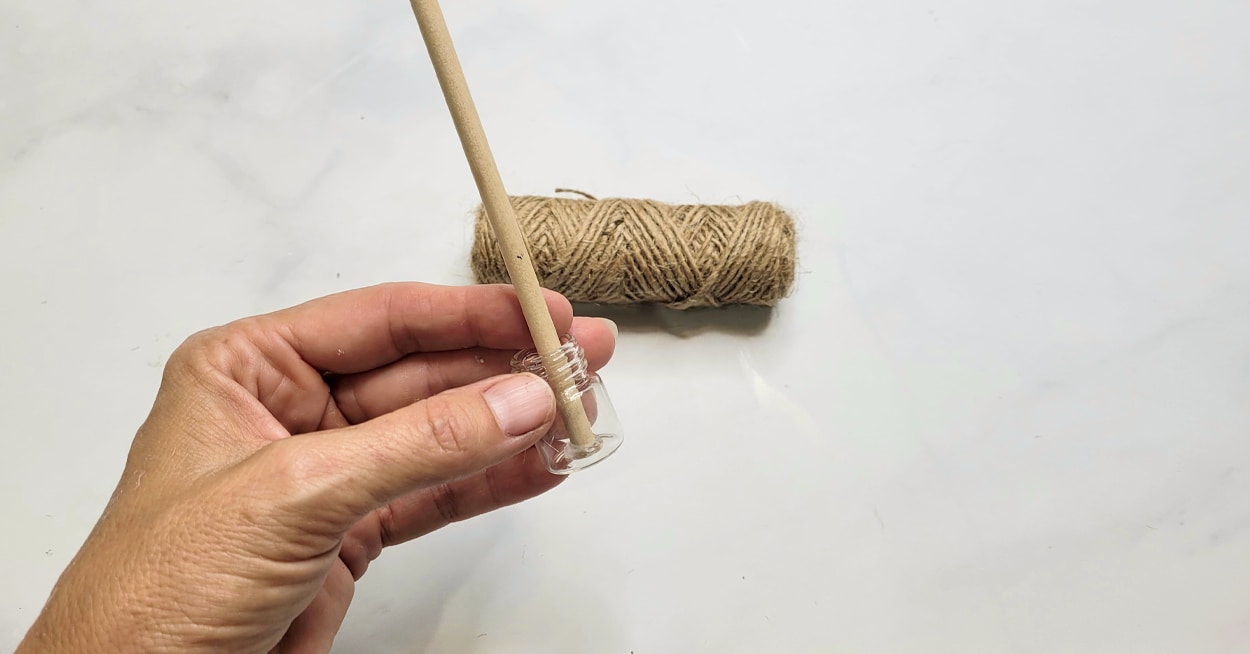 A hand holding a small glass container with a dowel rod in it and twine
