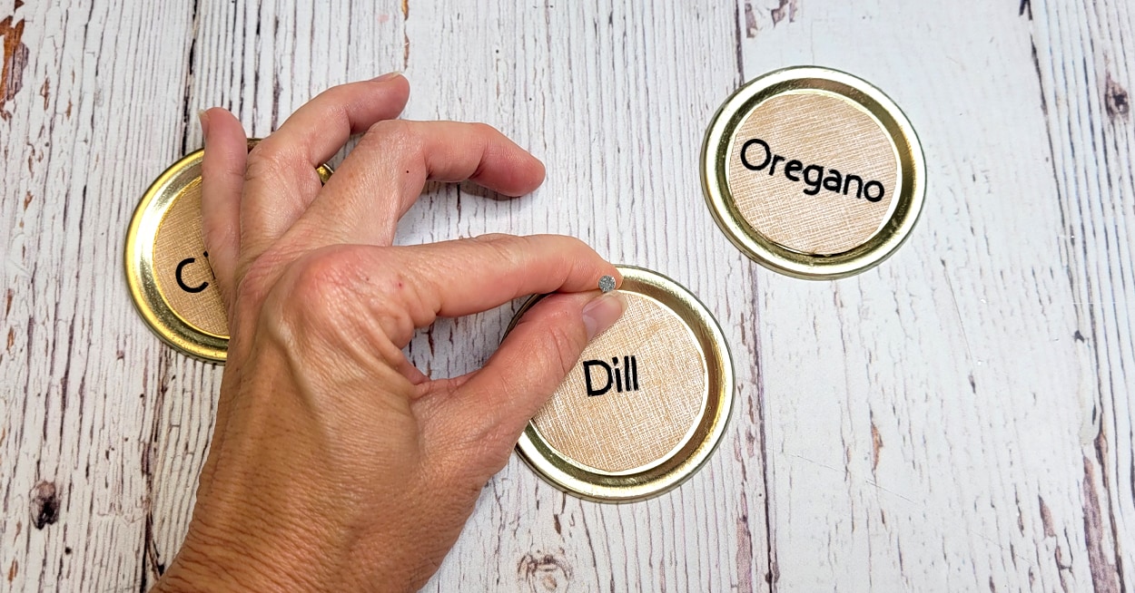 A hand holding a nail on a lid with cardstock and lettering on it.