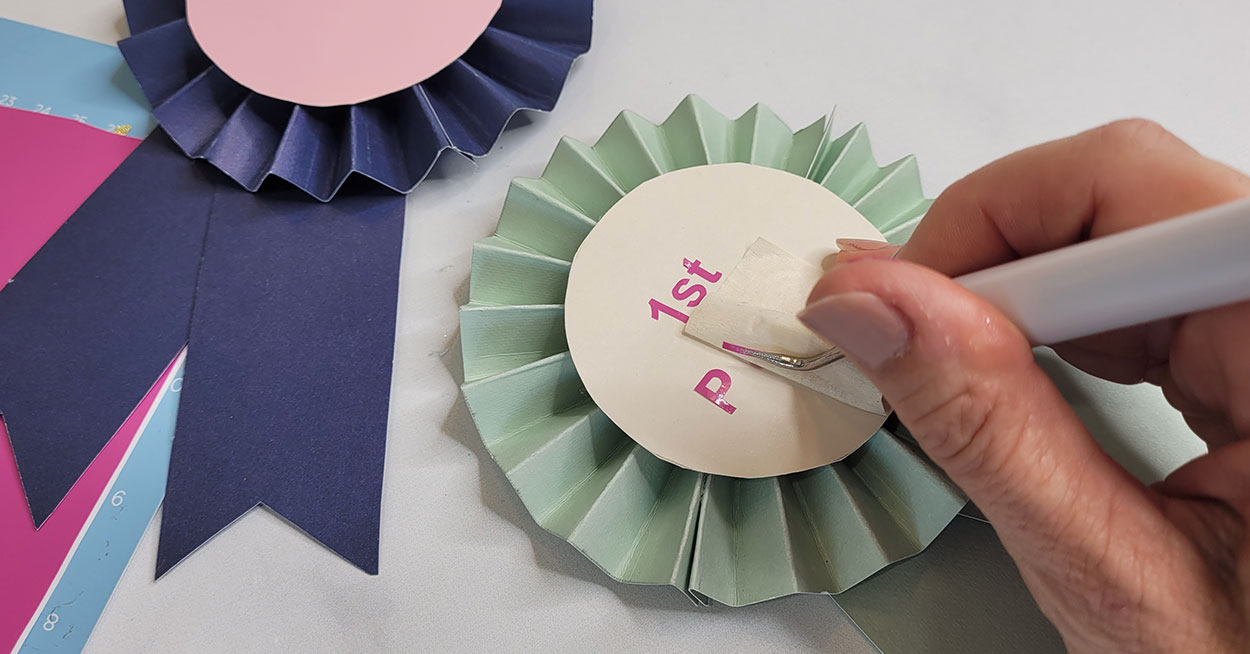 A hand holding a craft picker, vinyl lettering, and a paper award ribbon.